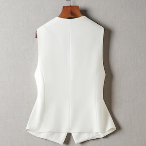 May include: A white sleeveless vest with a tailored design. The back view reveals a V-neck and a split hem. The vest is suspended on a wooden hanger against a neutral backdrop.