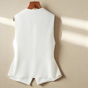 May include: A white sleeveless vest hanging on a wooden hanger. The vest features a V-neck and a tailored fit, with a split hem at the back. The fabric appears to be a smooth, woven material.
