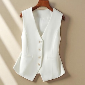 May include: A white vest with a V-neck design, tailored with three decorative buttons. The vest has two front pockets and is hanging on a wooden hanger. This garment is suitable for formal or business attire.