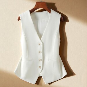 May include: A white waistcoat with a V-neck and four buttons. The waistcoat has two front pockets and is hanging on a wooden hanger. The fabric is smooth and the fit is tailored.