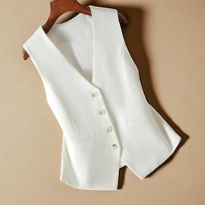 May include: A white, tailored waistcoat with a V-neck and four button closure. The waistcoat has two front pockets and is hanging on a wooden hanger. The garment is a formal style, against a neutral background.