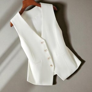 May include: A white waistcoat with a V-neck and button closure. The tailored vest features two mock pockets and hangs on a wooden hanger. The garment is made of a smooth, solid-coloured fabric.