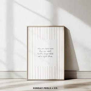 May include: A framed art print in neutral tones. The artwork has a white background with vertical beige stripes and a central white rectangle with handwritten text. The text reads: "when you have more than you need, build a longer table not a higher fence."