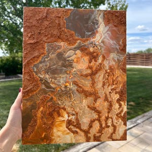Peut inclure: Œuvre d'art abstraite avec une surface texturée. L'œuvre présente un mélange de tons terreux, notamment des nuances de marron, d'orange et de crème, créant une sensation de profondeur. L'œuvre est tenue par une main, montrant sa taille et sa texture.