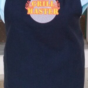 May include: Black apron with the words "GRILL MASTER" in orange and yellow, surrounded by a flame design. The apron has black straps and is worn over a light blue collared shirt.