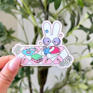 May include: A sticker featuring a white bunny wearing glasses and a pink dress, reading a book on a pink and white checkered blanket. A blue purse, books, and carrots are also on the blanket. Daisies and flowers surround the bunny.