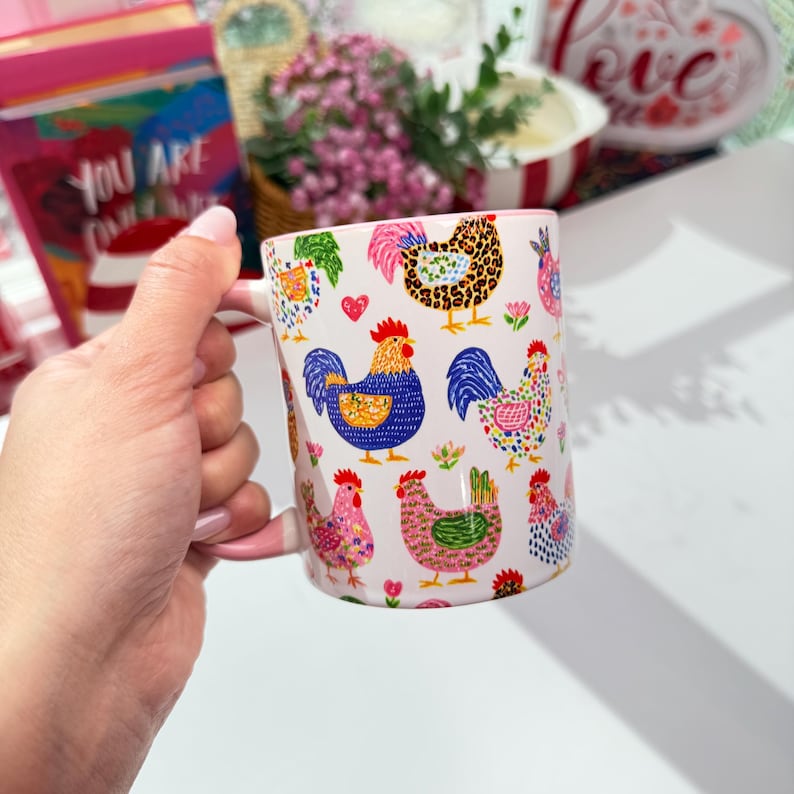A hand holds a white ceramic mug with a pink rim and handle, decorated with colorful cartoon chickens and roosters, casting a shadow on a white surface.