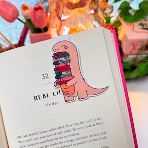 May include: A peach-colored dinosaur bookmark clipped onto an open book page. The dinosaur is illustrated with a smiling face and is holding a vertical stack of six books in various colors including red, green, and dark teal, with black outlines.