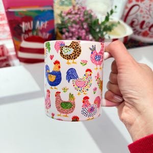 A hand holds a white ceramic mug with a pink rim and handle, decorated with colorful cartoon chickens and roosters, with a blurred red and white background.