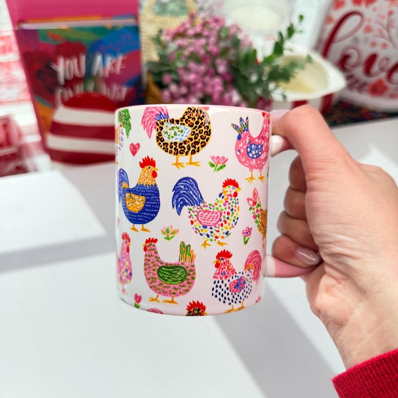 A hand holds a white ceramic mug with a pink rim and handle, decorated with colorful cartoon chickens and roosters, with a blurred red and white background.