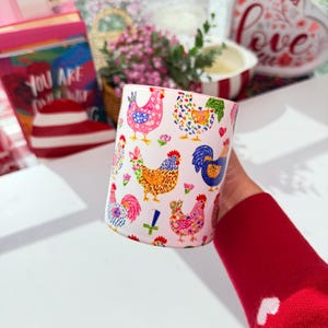 A hand in a red sleeve holds a white ceramic mug with a pink rim and handle, decorated with colorful cartoon chickens and roosters, seen from a side angle.