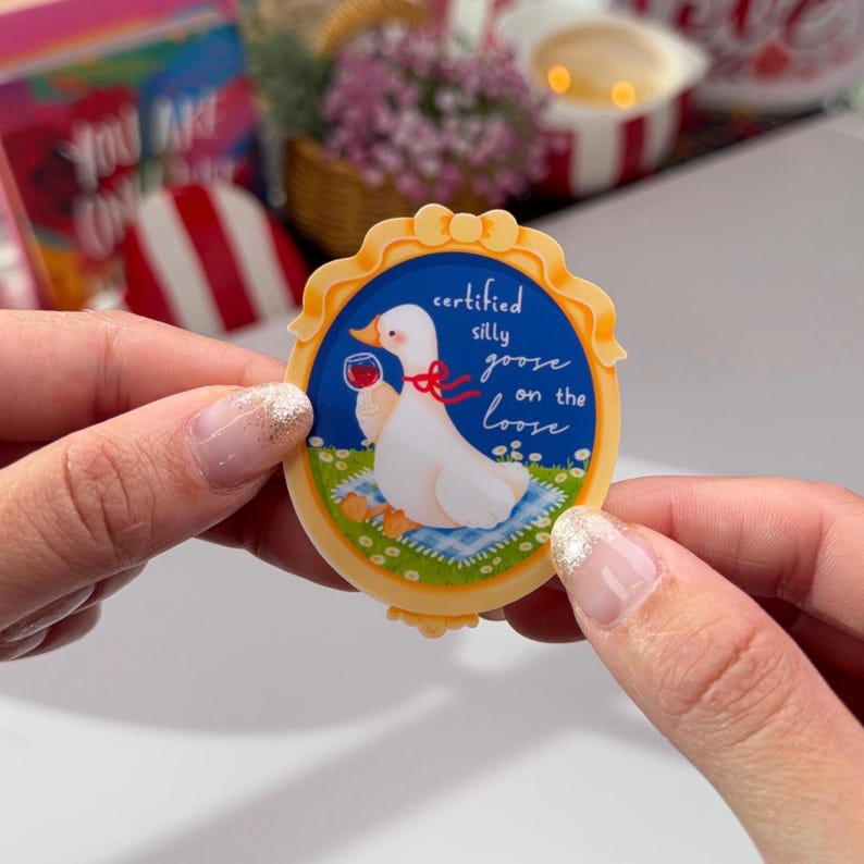 May include: A circular sticker featuring a cartoon goose with a glass of red wine. The goose is on a picnic blanket with flowers, and the text reads "certified silly goose on the loose". The sticker has a yellow border.