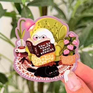 May include: A die-cut sticker featuring a white ghost with round glasses sitting in a green armchair, reading a book titled How to Haunt Your Enemies Vol. I. A black cat lies on a patterned rug near a cup of tea and a potted pink flower plant.