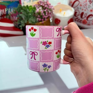 May include: A pink and white ceramic mug with a checkered pattern and floral designs. The mug features pink bows and various flowers, including red tulips and blue and purple flowers. The handle is also pink.
