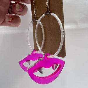 May include: A pair of hoop earrings with a pink acrylic charm shaped like a mouth with fangs. The hoops are made of white beads.