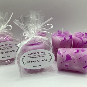 May include: Two clear cellophane bags with purple votive candles inside. The bags are tied with white ribbon and have a label that reads "Candles by Laura" and "Cherry Almond".