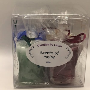 May include: A clear plastic box containing two scented candles. One candle is green, the other is burgundy. Each candle is wrapped in clear cellophane and tied with a ribbon. The label reads "Candles by Laura, Scents of Maine, USA."