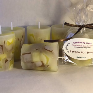May include: Four yellow candles with a brown and white design. The candles are packaged in a clear plastic bag with a brown ribbon and a label that reads "Candles by Laura", "Hand poured - One of a kind", "www.lstubbs.etsy.com", "Banana Nut Bread", and "Maine USA".