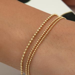 May include: A delicate gold-toned bracelet featuring a fine ball chain design. The jewellery piece consists of two parallel strands of small, uniform metallic spheres linked together, creating a simple and refined appearance on the wrist.