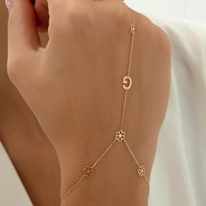 May include: A gold hand chain featuring a delicate link design with small flower-shaped charms and a single letter G charm. The jewelry piece connects from the wrist to a finger, showcasing a polished metallic finish and a fine chain construction.