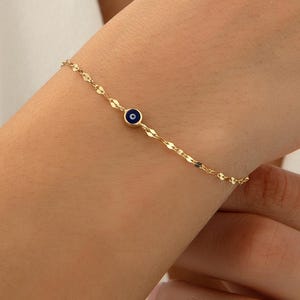 May include: A delicate gold chain bracelet featuring a single, small, circular evil eye charm. The charm consists of a dark blue center surrounded by a white ring and a gold border, set against a thin, reflective gold link chain.