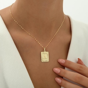 May include: A gold-toned necklace featuring a rectangular pendant with a textured border and the letter A embossed in the center. The pendant hangs from a delicate, shimmering chain that rests against the collarbone, creating a refined look.