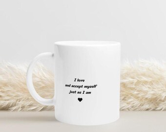 May include: White ceramic mug with a curved handle. The mug features the text "I love and accept myself just as I am" in black, with a black heart symbol below the text. The mug is set against a neutral background.