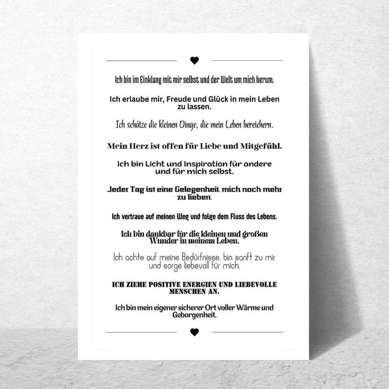 May include: A white rectangular print with black text. The text is in German and includes affirmations about self-love, gratitude, and attracting positive energy. A small black heart is at the top and bottom.