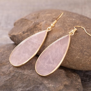 May include: A pair of teardrop-shaped earrings with a pale pink stone set in a gold-colored metal frame. The earrings are displayed on a gray stone surface, showcasing their delicate design and color.