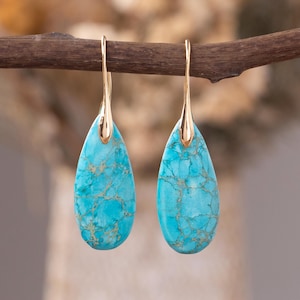 May include: A pair of gold-colored drop earrings with teardrop-shaped turquoise stones. The turquoise stones have a marbled pattern with gold-colored veins. The earrings are hanging from a brown branch.