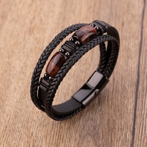 Puede incluir: Pulsera de cuero negro con bandas trenzadas y lisas. La pulsera presenta tres piedras rectangulares marrones y un cierre de metal negro. La pulsera se muestra sobre una superficie de madera.