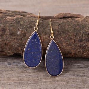 May include: A pair of teardrop-shaped earrings with a deep blue stone and gold-colored metal trim. The earrings are displayed against a textured wooden background, showcasing their elegant design and color contrast.