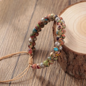 Peut inclure: Un bracelet de perles multicolores avec des perles rondes rouges, vertes, bleues et marron. Le bracelet comporte des accents métalliques argentés et est monté sur un cordon beige. Le bracelet est présenté sur une surface en bois.