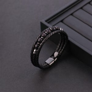 May include: Black leather bracelet with three strands, featuring dark lava stone beads and silver-tone accents. The bracelet has a magnetic clasp and a braided design, perfect for everyday wear.