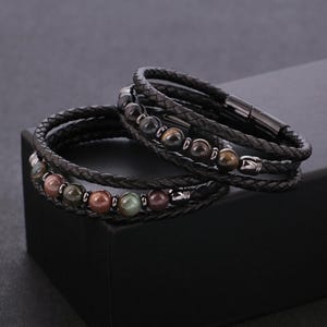 Könnte beinhalten: Zwei schwarze Lederarmbänder mit geflochtenen Bändern. Jedes Armband hat eine Reihe polierter, mehrfarbiger Edelsteinperlen. Die Armbänder werden auf einer schwarzen Oberfläche präsentiert, die ihr Design und ihre Textur hervorhebt.