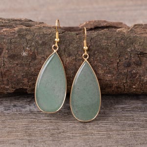 May include: A pair of teardrop-shaped earrings with a light green stone set in a gold-colored metal frame. The earrings hang from gold-colored hooks. The earrings are displayed on a wooden surface.