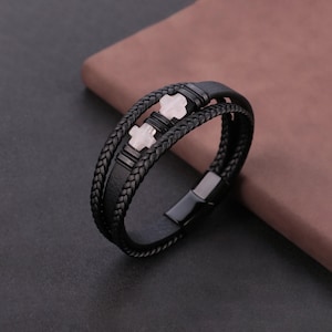 May include: Black leather bracelet with a braided design and a magnetic clasp. The bracelet features two light-colored cross-shaped accents. The bracelet is shown on a dark surface.