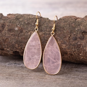 Pode incluir: Um par de brincos em forma de gota com uma pedra rosa pálido emoldurada em metal dourado. Os brincos pendem de ganchos dourados. O fundo é uma superfície de madeira.
