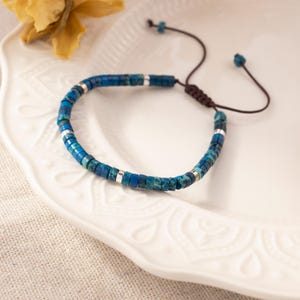 Könnte beinhalten: Verstellbares Armband mit blauen und silbernen Perlen. Das Armband besteht aus kleinen, quadratischen blauen Perlen mit silbernen Akzenten. Die verstellbare Kordel ist braun. Das Armband wird auf einem weißen, dekorativen Teller präsentiert.