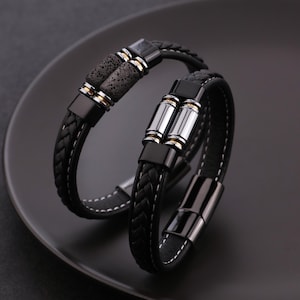 Puede incluir: Dos pulseras de cuero negro, una con cuero trenzado y cuentas de piedra de lava gris oscuro, la otra con cuero liso y detalles cilíndricos plateados. Ambas tienen cierres magnéticos negros y costuras blancas.