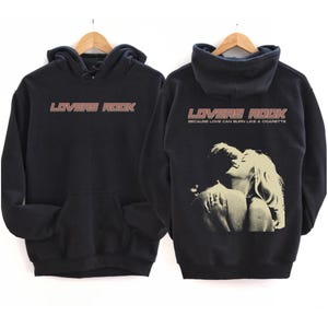 May include: Black hoodie with a graphic print of a couple embracing. The text "LOVERS ROCK" is printed on the front of the hoodie in a pink and white font. The text "BECAUSE LOVE CAN BURN LIKE A CIGARETTE" is printed on the back of the hoodie in a smaller font.