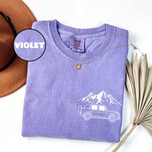 May include: Violet Comfort Colors t-shirt with a white graphic of a truck and mountain range. The shirt has a crew neck and short sleeves. A gold necklace with a pendant is draped on the shirt.