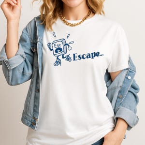 May include: White t-shirt with a blue cartoon graphic of an "Esc" key running, accompanied by the word "Escape..." in blue. The shirt is made of a soft material, suitable for casual wear.