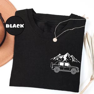 May include: Black t-shirt with a white graphic of a truck and mountain range. A silver necklace with a circular pendant rests on the shirt. The word "BLACK" is in a black circle in the upper left corner.