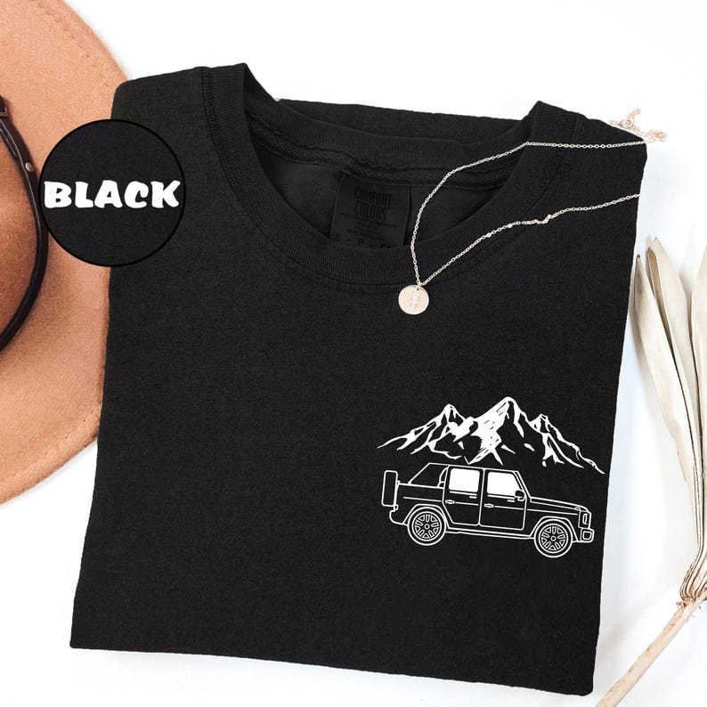May include: Black t-shirt with a white graphic of a truck and mountain range. A silver necklace with a circular pendant rests on the shirt. The word "BLACK" is in a black circle in the upper left corner.