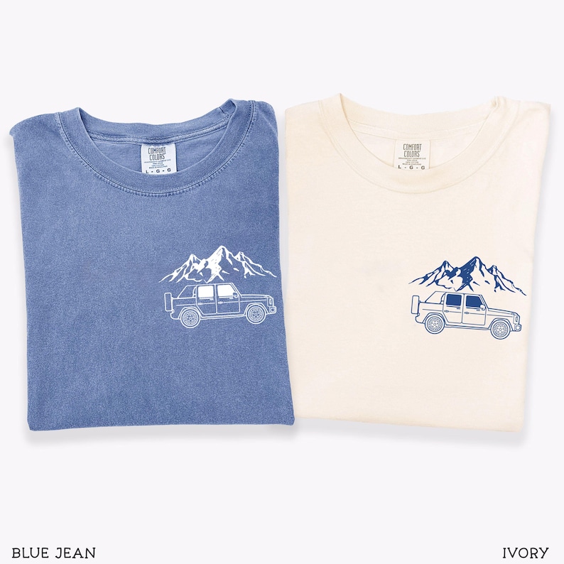 May include: Two t-shirts, one blue and one ivory, each with a mountain and truck graphic. The blue shirt has a white graphic, while the ivory shirt has a navy graphic. The shirts are folded and have a crew neck.