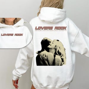 May include: White hoodie with a graphic print of a couple embracing. The text "LOVERS ROCK" is printed in brown on the front of the hoodie. The text "BECAUSE LOVE CAN BURN LIKE A CIGARETTE" is printed in brown below the graphic.