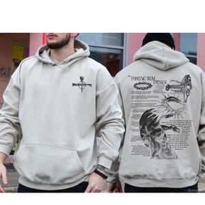 May include: Beige hoodie with a front pocket and a drawstring hood. The front features a black graphic of a cross with nails. The back displays the text "FORGIVE THEM FATHER..." along with a detailed illustration and script.