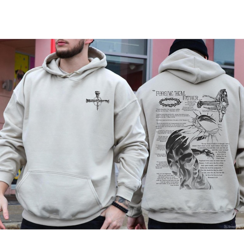May include: Beige hoodie with a front pocket and a drawstring hood. The front features a black graphic of a cross with nails. The back displays the text "FORGIVE THEM FATHER..." along with a detailed illustration and script.