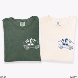 May include: Two folded t-shirts, one moss green and one ivory. Each shirt features a mountain and vehicle graphic on the front. The moss green shirt has a white graphic, while the ivory shirt has a blue graphic. Both shirts have a crew neck.